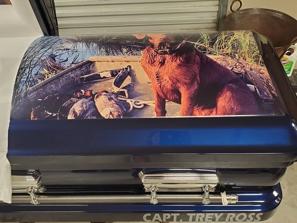 Navy casket topped with a duck boat photo of a retriever on the hunt, lettered Capt. Trey Ross