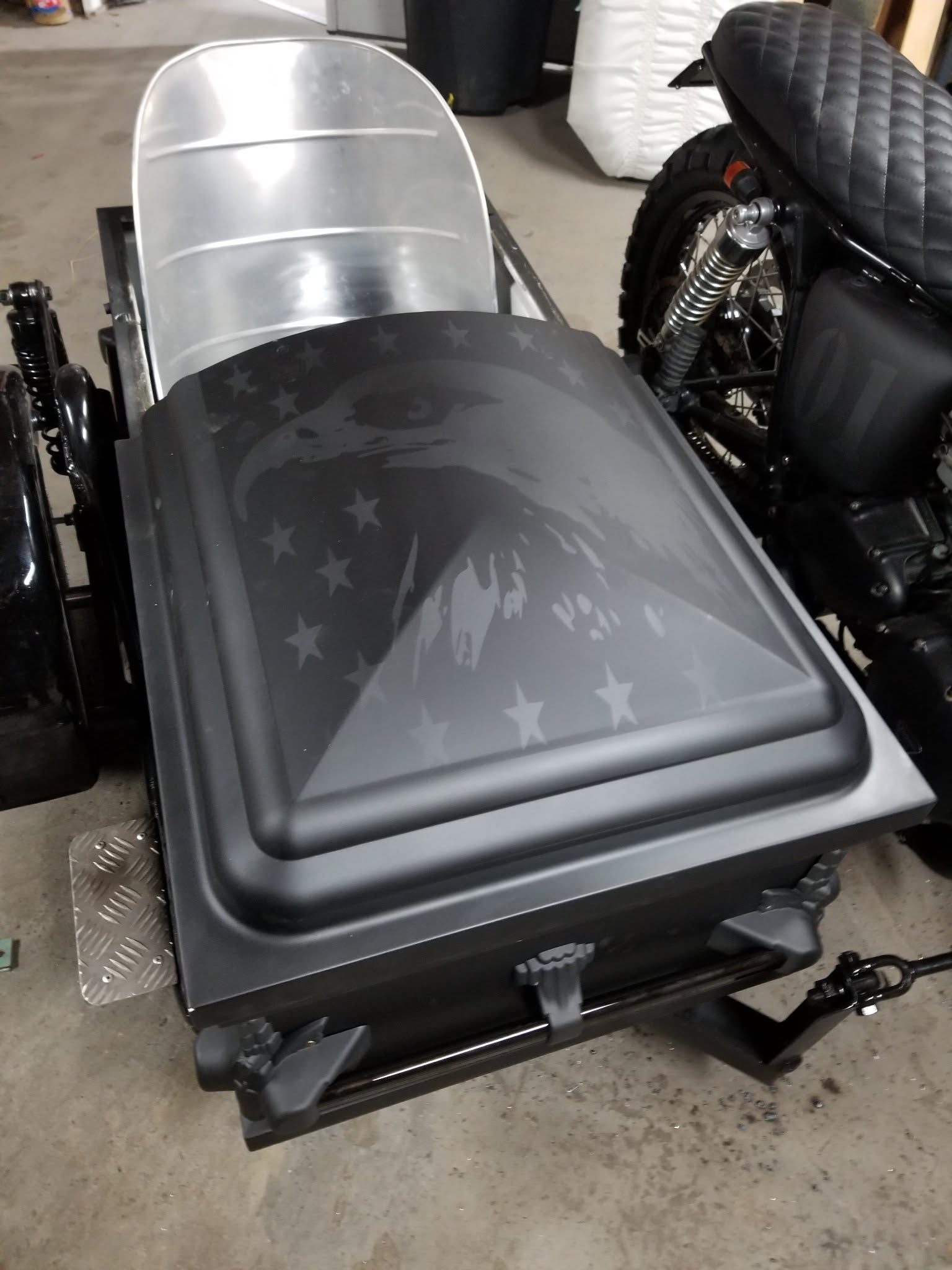Matte black casket lid wrapped with a bald eagle and stars, loaded on a motorcycle hearse trailer
