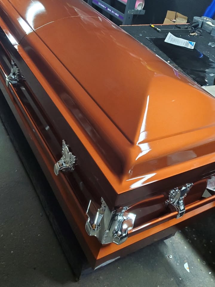 Candy orange metallic casket with ornate chrome corner handles resting under shop lights