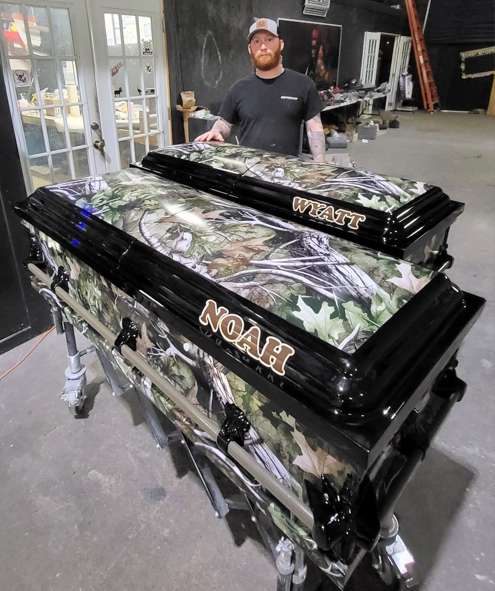 Leafy woodland camo casket lettered NOAH and WYATT with black trim, shown beside a SoulShine team member