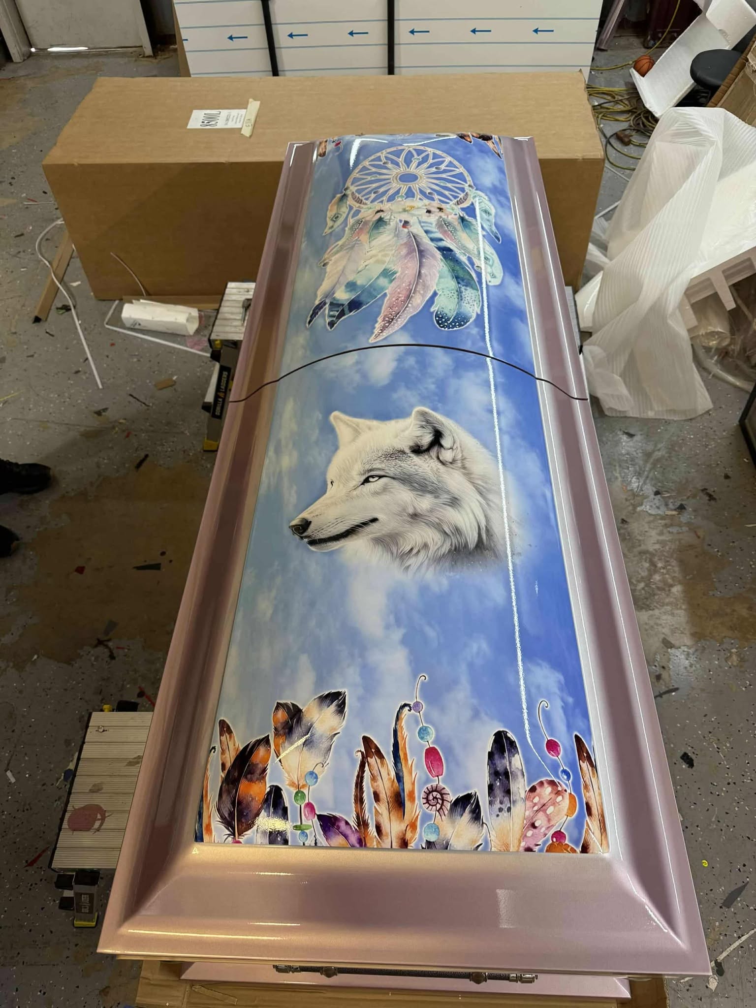 Rose pink casket topped with a white wolf, dreamcatcher and feather artwork on a blue sky