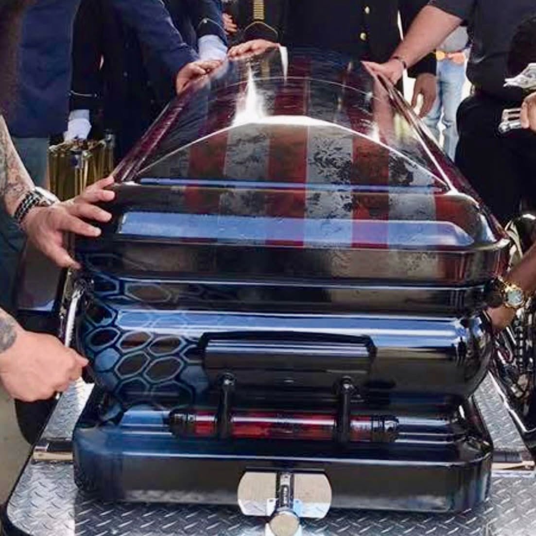Stars and stripes flag wrap over a glossy black casket carried by pallbearers at a graveside service