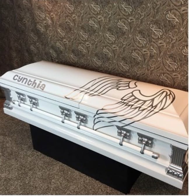 Pearl white casket scripted with cynthia beside a large outlined angel wing and nickel handles