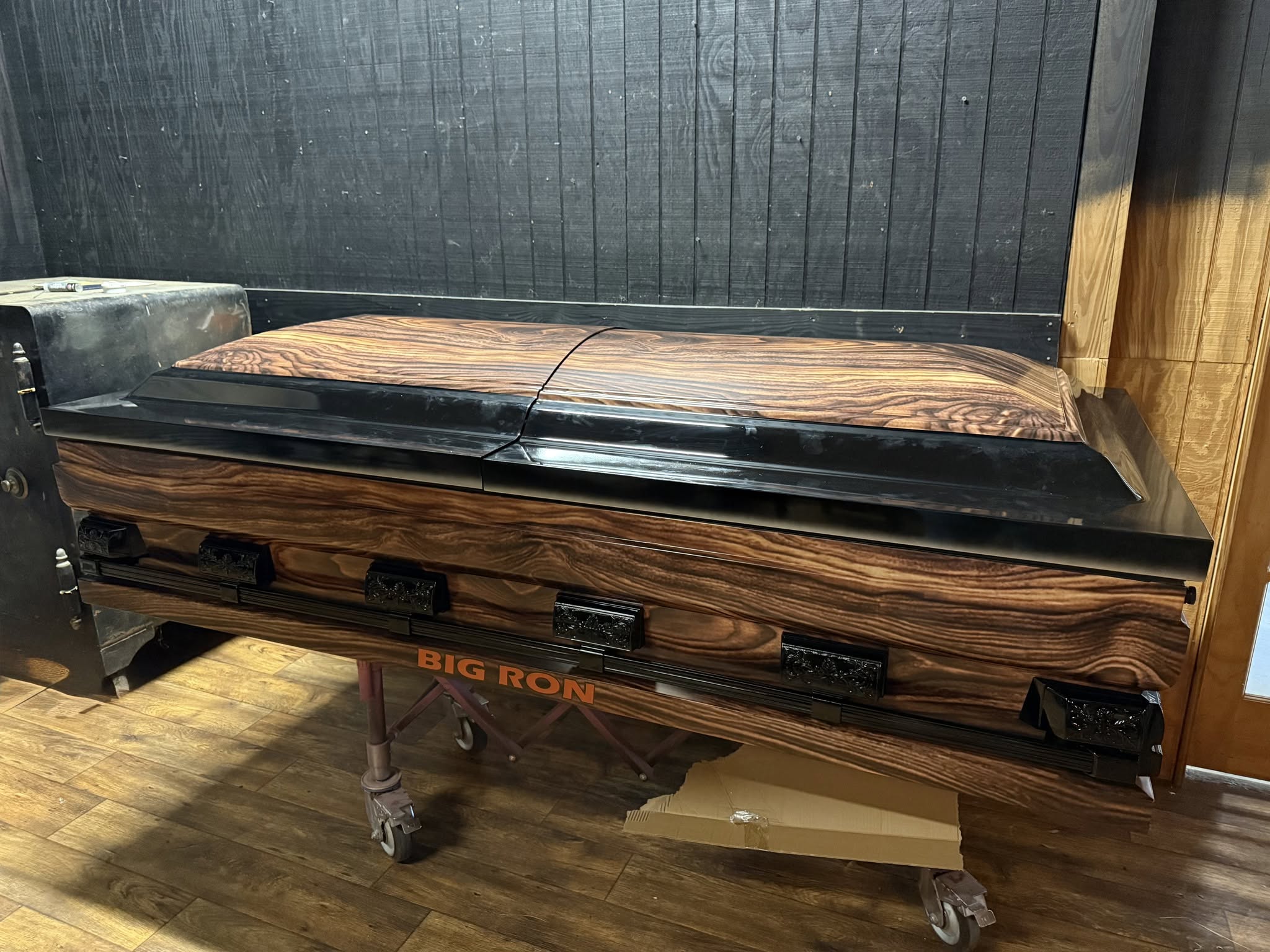 Extra wide wood grain casket with black trim resting on a church truck lettered BIG RON