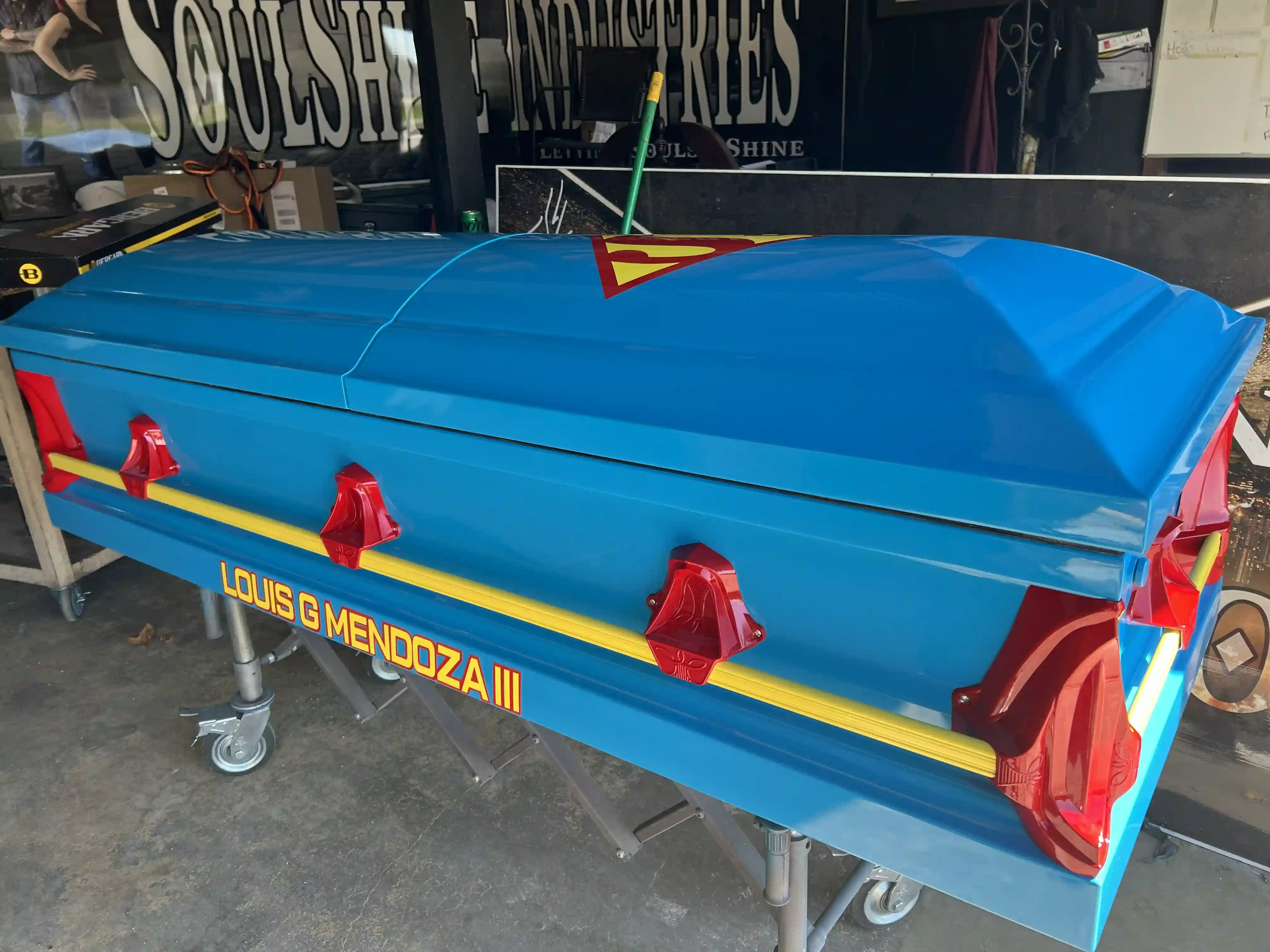Angled view of a bright blue Superman child casket with red corner handles and a gold rail